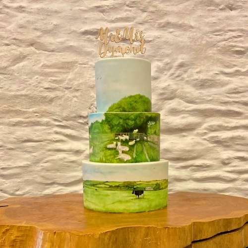 Three tier wedding cake with hand painted Cornish country side design, displayed at Trenderway Barns Cornwall