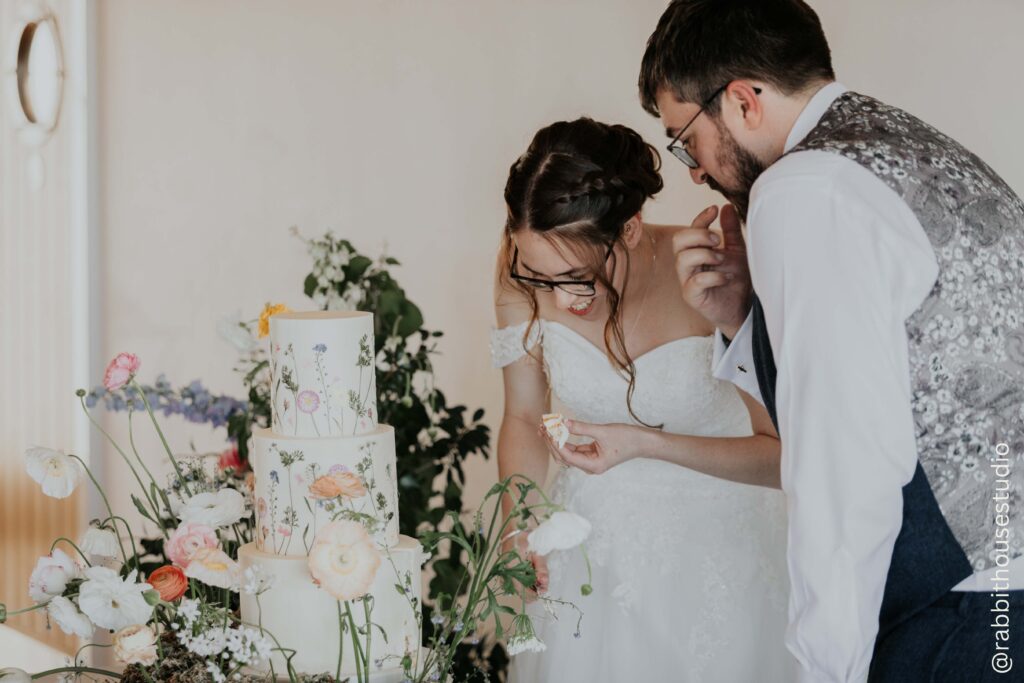 My couple cutting and tasting their wedding cake at The Green wedding venue. The cake is deocrated with pressed edible flowers to create a meadow feel cake design.